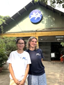 An image of two young female volunteers pictured in front of the Friends-International logo in the courtyard of the F3 center in Phnom Penh, Cambodia